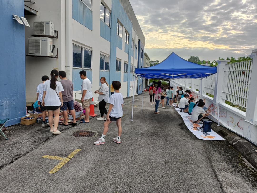 Volunteers painting the Celebrate the Beauty of Innocence mural along the white boundary wall of the Kiwanis Down Syndrome Foundation - National Centre (Malaysia), with paint pots and brushes laid out beside them.