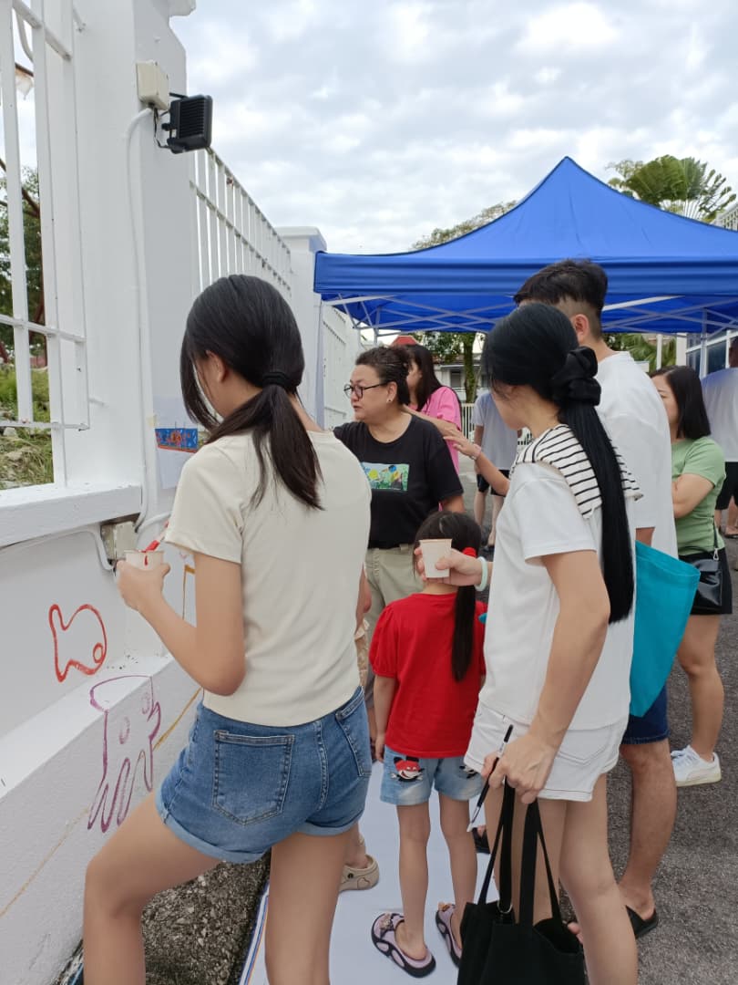 A volunteer painting a red fish detail on the mural wall during the JKM Petaling volunteer morning, holding a paper cup of paint.
